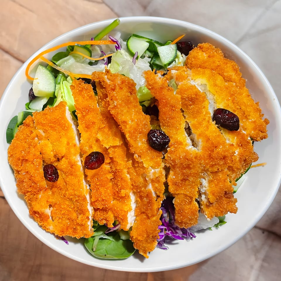 Crispy fried chicken cutlets with dried cranberries on a fresh green salad in a white bowl.