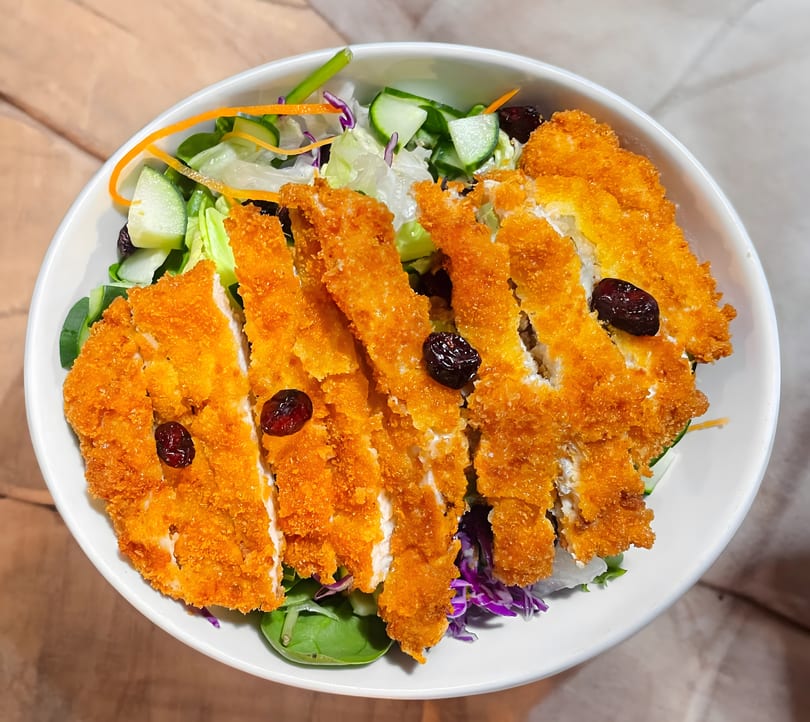 Crispy fried chicken cutlets with dried cranberries on a fresh green salad in a white bowl.