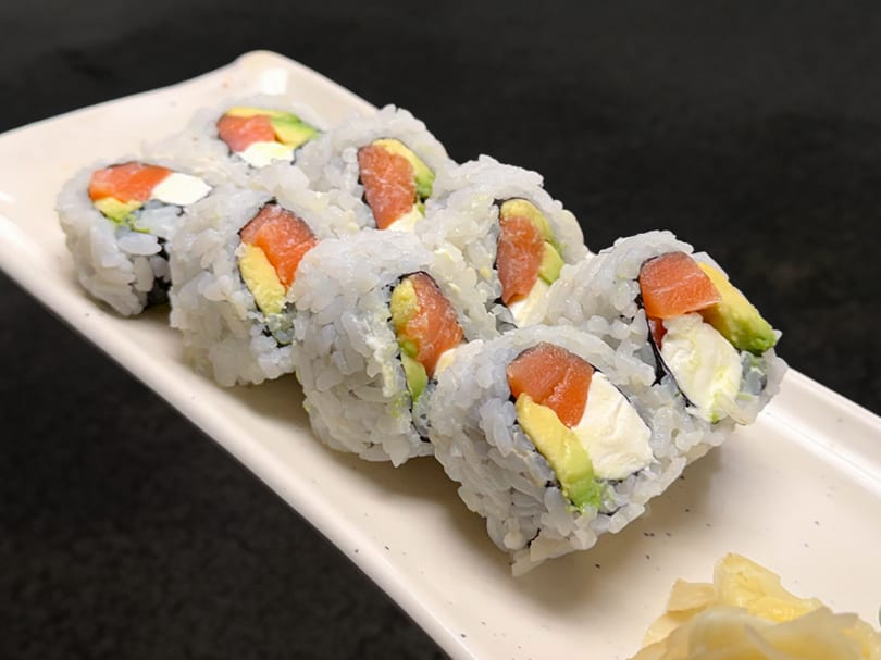 Six sushi rolls with salmon, avocado, and cream cheese on a white rectangular plate.