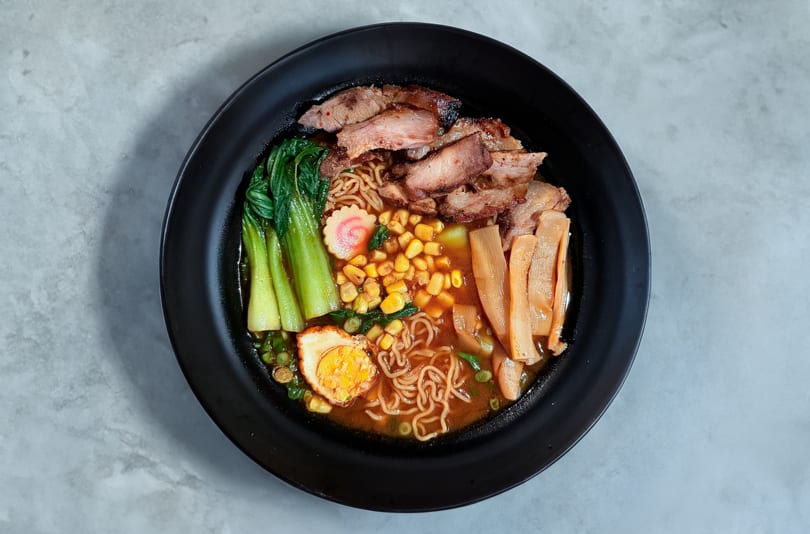 A black bowl of ramen with pork, bok choy, corn, bamboo shoots, and a soft-boiled egg.