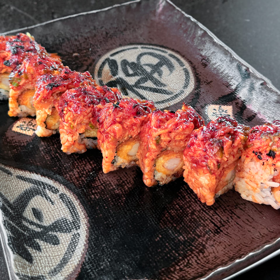 Spicy sushi roll with red topping and tempura shrimp on a dark patterned plate.
