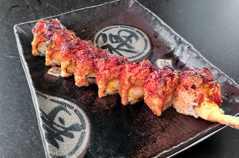Spicy sushi roll with red topping and tempura shrimp on a dark patterned plate.