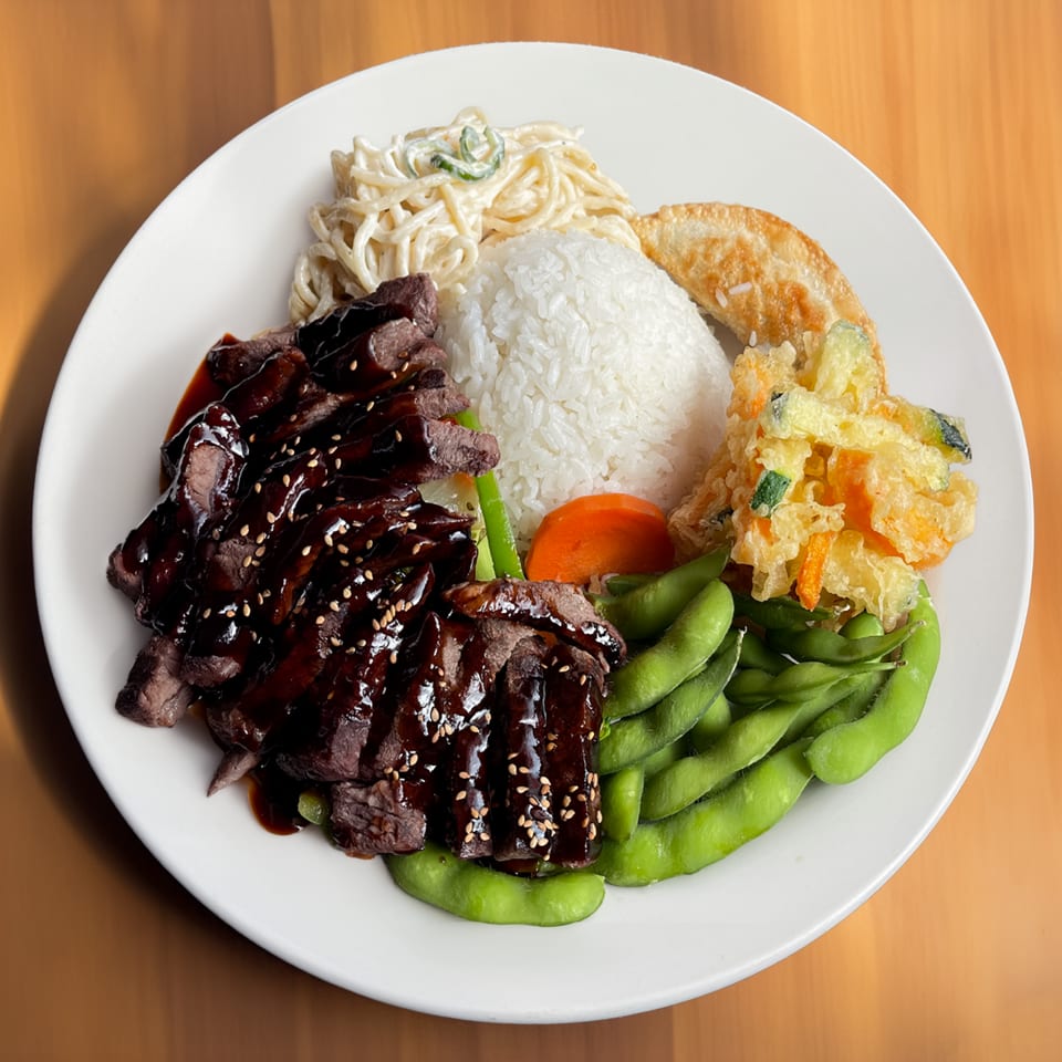 Teriyaki beef with rice, noodles, edamame, tempura vegetables, and a fried dumpling on a white plate.