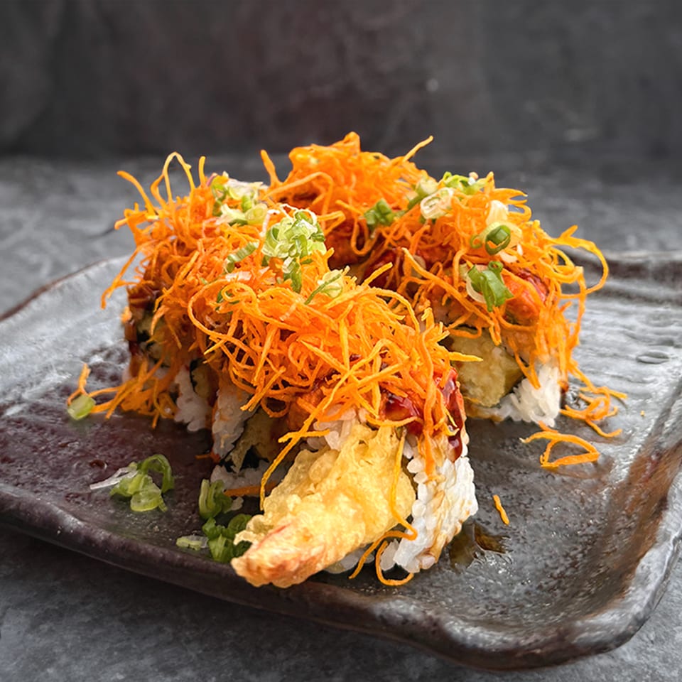 Sushi rolls topped with crispy orange strands and green onions on a dark rectangular plate.