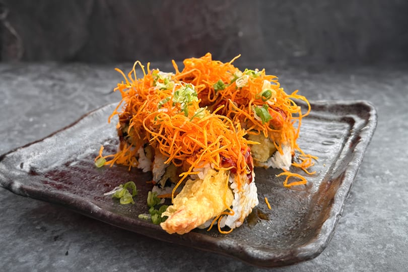 Sushi rolls topped with crispy orange strands and green onions on a dark rectangular plate.
